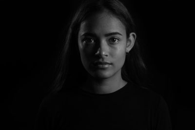 A portrait of an aboriginal girl in her late teens, wearing a black t-shirt. She is facing the camera and has long hair that reaches down to her shoulders. The lighting is dark with high contrast and the background color should be a deep black. This photo was taken using a Canon EOS R5 camera and Canon RF24-70mm f/8 IS USM lens. It is a black and white, high resolution photograph in the style of a dark, high contrast portrait. --ar 128:85