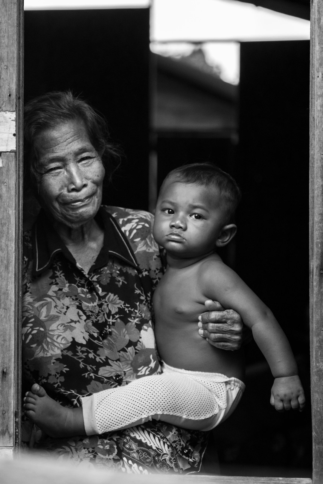 A black and white photograph shows an old Indonesian woman with her arms around the shoulders of a very young boy. He is wearing knitted leg warmers as they look out from behind an open wooden door towards the camera. The scene has a strong composition on the left side with lots of negative space on either side. It is a high-contrast photograph with sharp focus, in the style of documentary photography. –ar 85:128