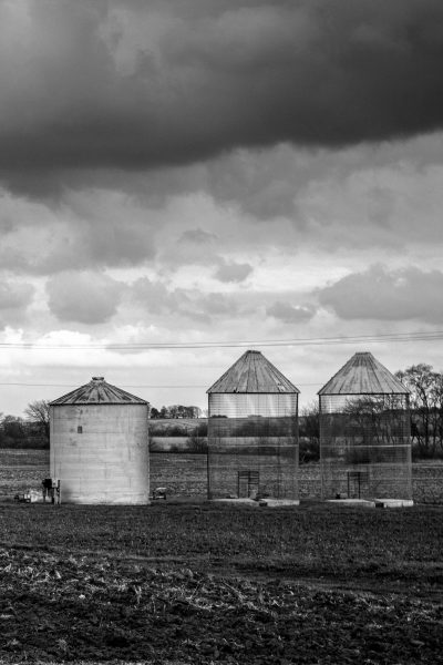 black and white photography of three modern silos in the middle distance, surrounded by farmland, with dark clouds overhead. The photograph is in the style of [Ansel Adams](https://goo.gl/search?artist%20Ansel%20Adams). --ar 85:128