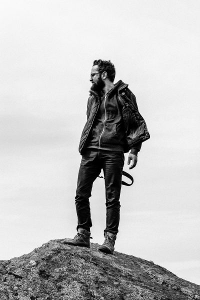 Black and white photography of stylish man standing on top rock, wearing leather jacket, jeans pants and boots shoes, portrait photography, --ar 85:128