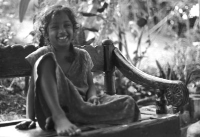 A happy young Indian girl sitting on an old wooden bench in the garden of her house, smiling at the camera. She is wearing traditional  and is barefoot. The scene has lots of daylight and there is a water pump behind it. Shot with Kodak film using a Leica M3 camera. Black and white photo in the style of street photographers of the era. --ar 16:11