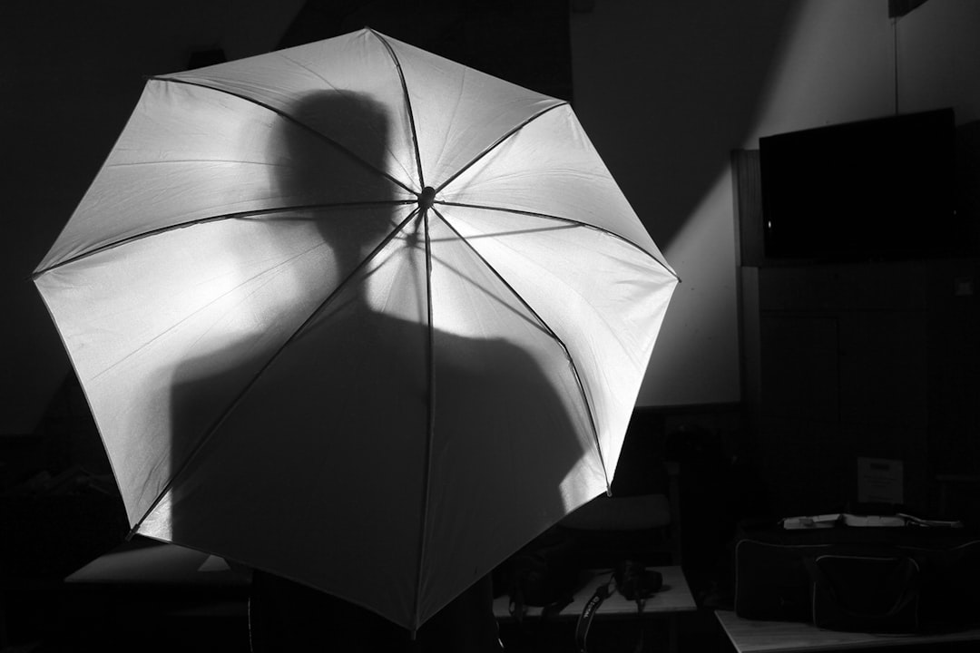 A black and white photo of an open umbrella casting a shadow on the wall, taken from behind the person holding it. The background is dark with some light coming through small windows, creating subtle shadows around the edges of the umbrella’s silhouette. In front there’s another object which adds to the composition. This scene has a monochromatic feel with high contrast between bright highlights and deep blacks, in the style of a minimalist photographer. –ar 128:85