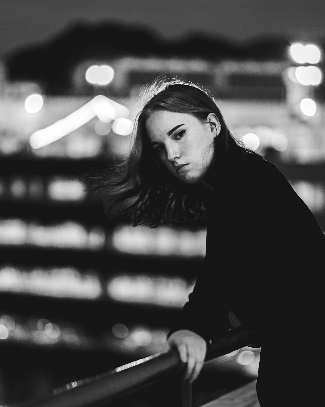 A black and white portrait of a young woman leaning over the edge of a bridge, city lights in the background, shot with a Fujifilm X-H2S and Canon K35 prime lens at 70mm in the style of anonymous. –ar 51:64