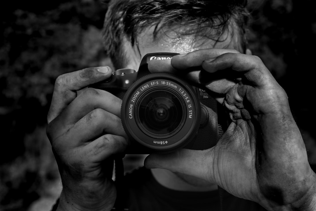 A black and white photo of an man holding the camera in front his face, taking pictures with it. The focus is on one hand that holds canon dslr 5d mark iv lens. He has short hair and facial stubble. In background we can see nature, trees and rocks. It’s late evening and there was light coming from left side of frame. Black and White photography, high contrast, grainy image texture –ar 128:85