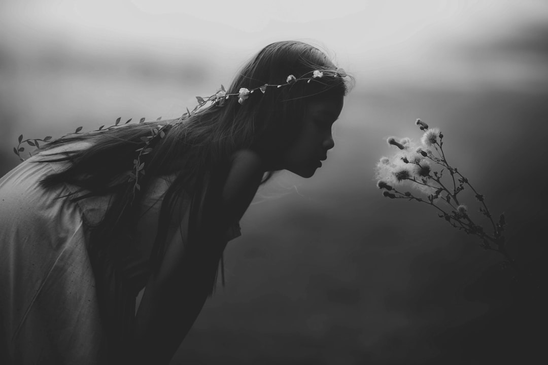 black and white photo of an angelic woman with long hair, wearing flower crown standing in the foggy field holding wild flowers while she is nose close to one single flower, her back facing camera, low angle shot, blurry background, cinematic, moody, sad, melancholic, ethereal, dreamy, –ar 128:85