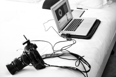A black and white photo of an open laptop on the edge of a hotel bed, with cables leading to a camera. The background is plain white, focusing attention on the equipment laid out for use in commercial photography. --ar 128:85