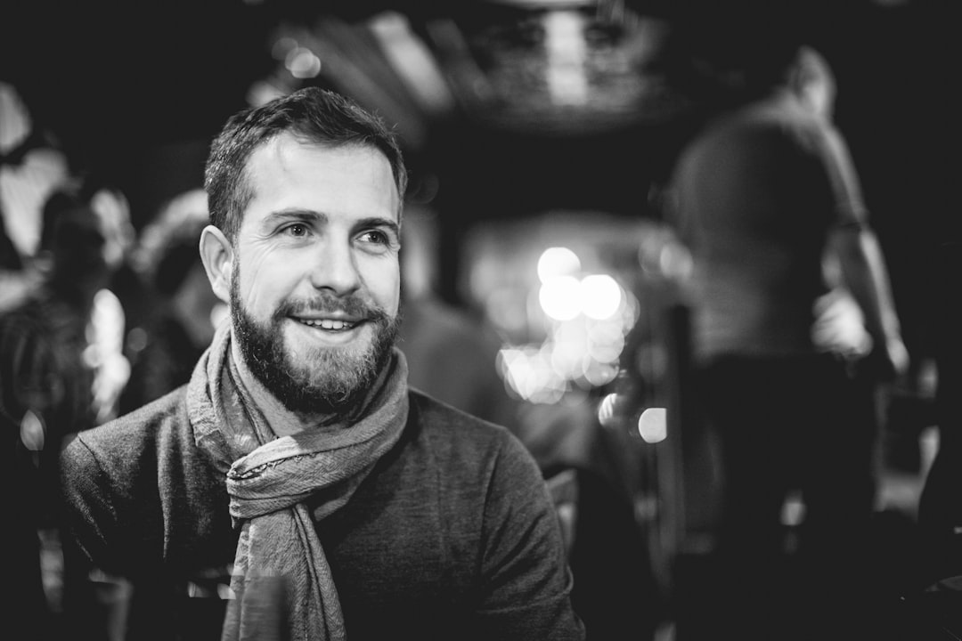 A handsome man in his thirties with short hair and beard, smiling at the camera while sitting on stage of an open-air bar during nighttime. The background is blurred to emphasize him. He’s wearing a scarf around his neck, dressed casually for fall weather. In sharp focus in the foreground, with a depth of field and bokeh effect, this black and white photography is in the style of a Leica Q2 Monochrom photograph. –ar 128:85