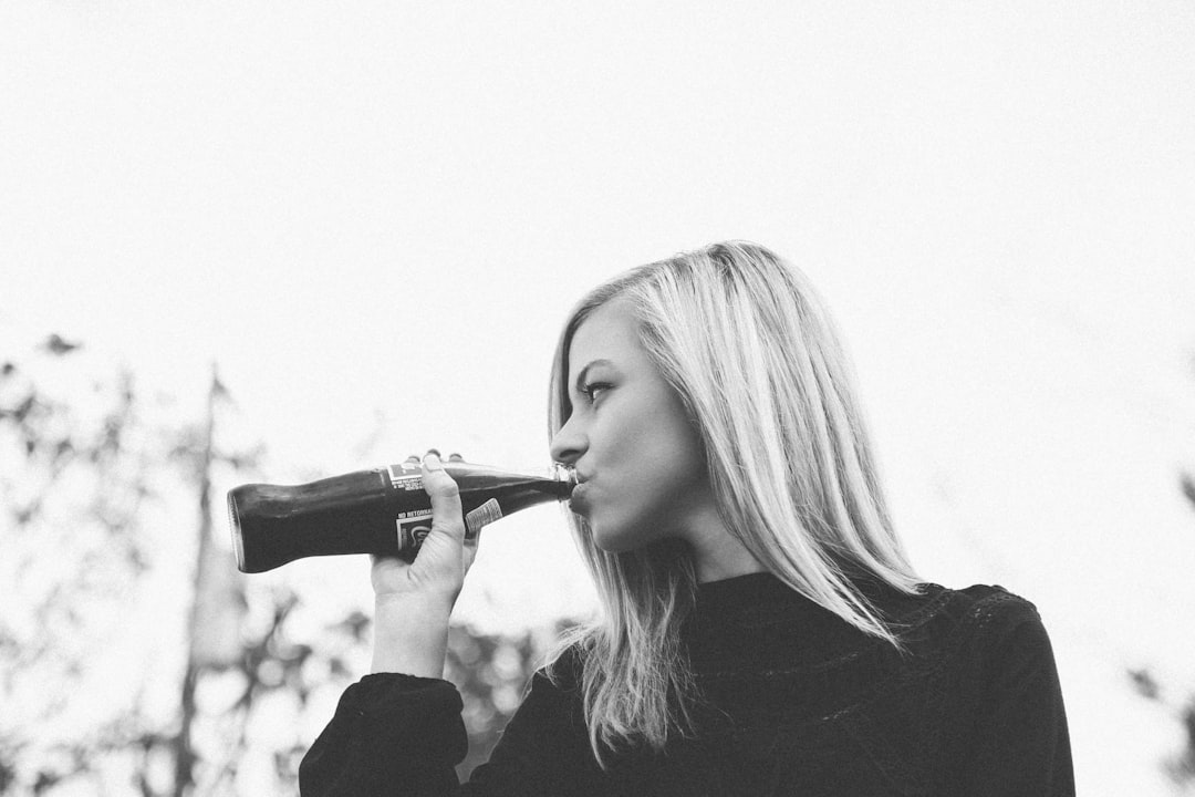 Black and white photo of a blonde woman drinking from a bottle, in profile view, in an outdoor setting, from a low angle, shot on a Hasselblad medium format camera with Kodak Portra film, in the style of minimalistic and candid photography. The photograph captures joyful moments and candid moments in a vintage black and white style. –ar 128:85