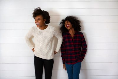 Two happy black women standing against a white wall. One woman is wearing a red plaid flannel shirt and jeans with curly hair laughing out loud, the other wears a cream sweater and blue jeans laughing out of love. Studio photography with a white background, in the style of anonymous. --ar 128:85