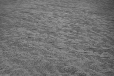 Black and white photograph of the sand texture, taken from above. The grainy texture has high contrast in the style of Leica M6 with SummiluxM cooke panchros lens at f/2.8. Natural light was used with professional color grading and soft shadows. The image has clean, sharp focus with a cinematic style of photography. --ar 128:85