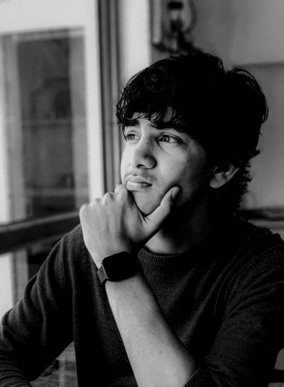 A young man with his hand on his chin, wearing an apple watch and a black sweater, is sitting in front of a table looking out at something outside. He has black hair and brown eyes. The image is in monochrome with a blurred background and is in the style of a Leica Q2 Monochrom. --ar 93:128