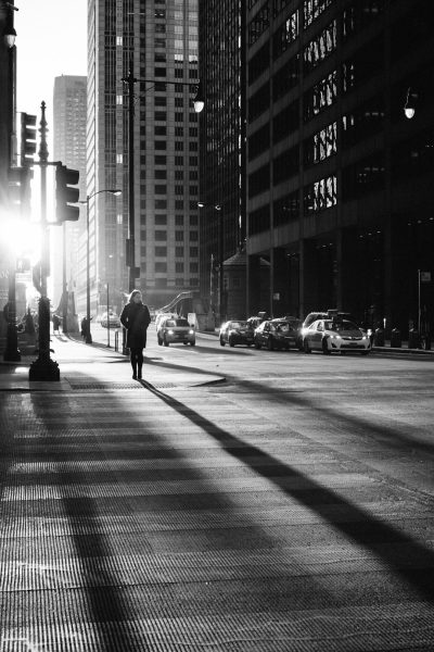 black and white street photography of a man walking in the streets of Chicago, early morning sun rays, skyscrapers, cars driving in the style of, shot on Leica M6 with f/8 lens --ar 85:128