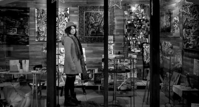 A black and white photo shows an art gallery inside a store. A woman stands in front of some paintings on display. She has short hair and is wearing a coat with a scarf around her neck and a hat. The walls have various works of art hanging. There is a glass window that reflects another street view outside. The photo has the style of Hasselblad X2D with a cinematic still similar to a [Wes Anderson](https://goo.gl/search?artist%20Wes%20Anderson) movie. It was taken at f/50mm and has a highly detailed background. --ar 128:69