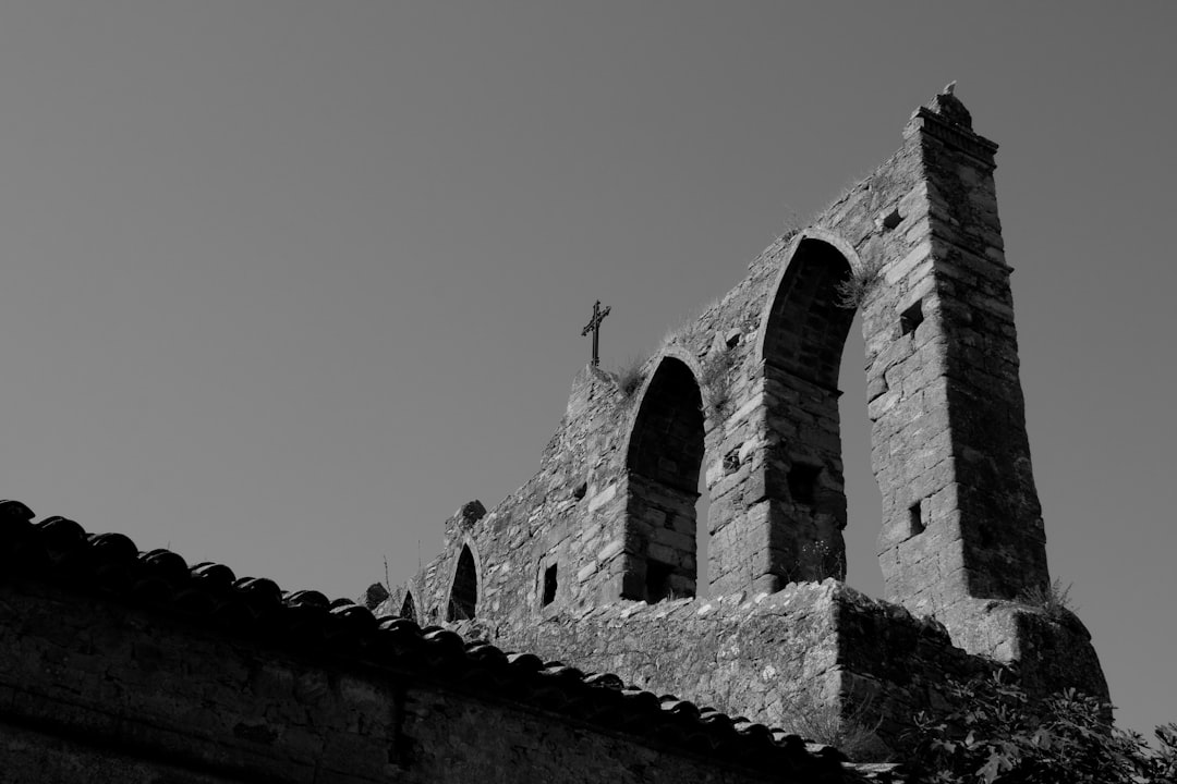 Black and white photography of an old church with a top roof, stone walls with arched windows and arches containing a cross, Spanish landscape on a sunny day with a minimalist style and negative space. –ar 128:85