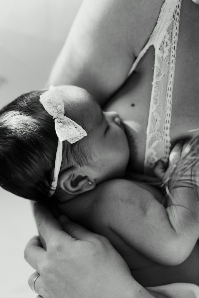 Close up of newborn baby girl being held in the style of her mother's torso, the little one's hand is on her mum's belly and she has a ribbon in her hair, baby lies back to hug mum, she wears a white lace top, black & grey photography, minimalism, in the style of Scandinavian and minimalist photography, natural light, high key, Nikon D850. --ar 85:128