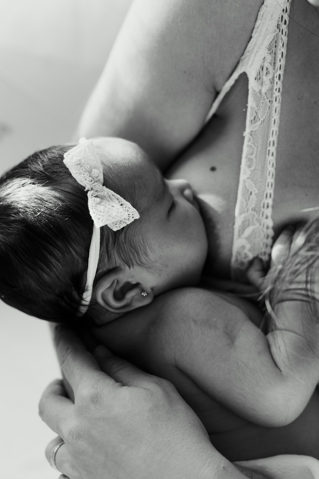 Close up of newborn baby girl being held in the style of her mother’s torso, the little one’s hand is on her mum’s belly and she has a ribbon in her hair, baby lies back to hug mum, she wears a white lace top, black & grey photography, minimalism, in the style of Scandinavian and minimalist photography, natural light, high key, Nikon D850. –ar 85:128