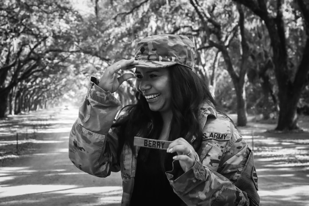 A black and white photo of a smiling woman in military fatigues with the word “Bidence” on her jacket, wearing a cap covering her hair, standing under oak trees at a Louisiana state park, pointing to the camera while showing off her name tag. She is holding her hand over her face smiling. –ar 128:85