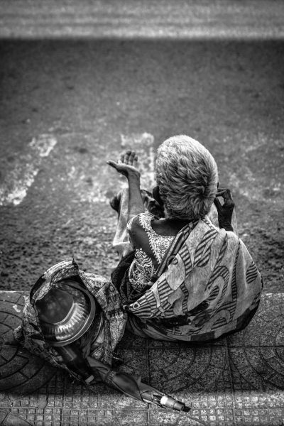 An old woman sitting on the ground, holding an iron pot and plate in her hands while she is performing alchemy magic to calm down someone who has fallen into Kailash MSkip. On the road of Mumbai, black and white, street photography in the style of high contrast, hyper realistic, wide angle, award winning photography in the style of super resolution, super sharp. --ar 85:128