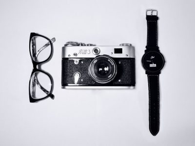 Black and white photography of a watch, camera, and glasses on a white background. The photograph is in the style of minimal editing of the original text. It captures everyday objects arranged on a plain surface. --ar 4:3