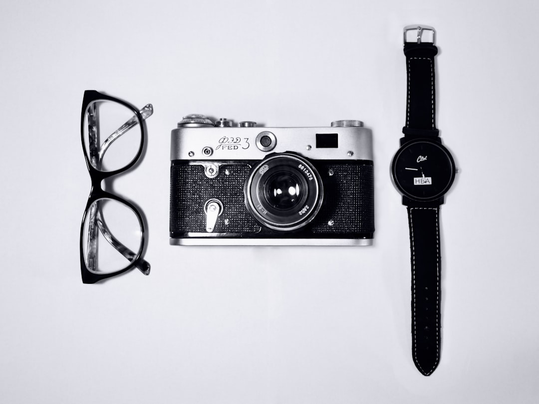 Black and white photography of a watch, camera, and glasses on a white background. The photograph is in the style of minimal editing of the original text. It captures everyday objects arranged on a plain surface. –ar 4:3