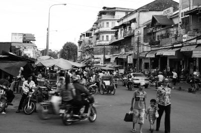 Black and white street photography of the city, busy streets with many people walking around in their daily lives, cars passing by on two sides, food stalls selling typical Vietnamese delicacies, motorbikes zooming past in all directions. The buildings along both sides of roads are old-style colonial architecture with flat roofs and black tiles. A group of children play happily at one corner while others walk to work or school. The overall mood is lively yet peaceful, capturing everyday life in Hanoi's Old Town area in the style of typical street photography. --ar 128:85