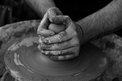 A black and white photo of hands holding clay on an archaic potter's wheel, focusing on the intricate details in the hands and clay textures. The background is blurred to emphasize the focus on sculpting the clay, creating a sense of artistry and craftsmanship. --ar 128:85