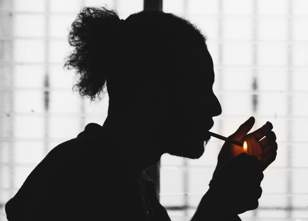 Silhouette of a black man lighting a cigarette with a match, in profile view, indoors, against a white background. The photograph was taken using a Sony Alpha A7 III with an aperture setting of f/28 to capture the details in his face. He has curly hair tied back in a ponytail, wearing a hoodie and jeans. The scene exudes an urban style, and there is no text or other elements present on it. –ar 32:23