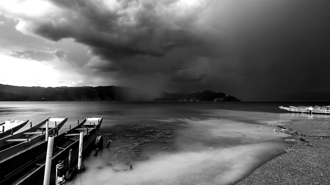 Black and white photography of the storm clouds over Lake Ripple Taf raging on. The pier is still there, but empty. There’s some boats scattered around. In the background you can see mountains far away from the lake with green vegetation. It was taken in the style of [Miki Asai](https://goo.gl/search?artist%20Miki%20Asai). High contrast. –ar 16:9