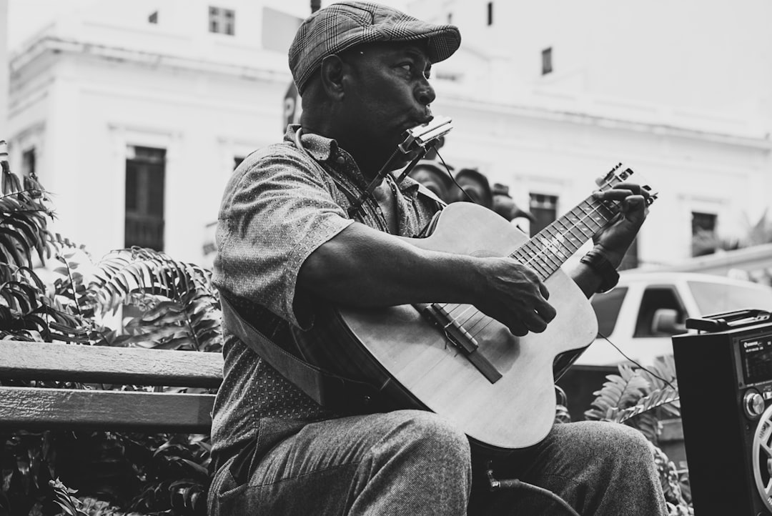 Black and white street photography of an Afro Latino man playing guitar in the streets of Old San Juan, Caribbean city with colonial architecture. The white buildings have lush greenery. 35mm film grain captures the scene in the style of a Leica M6 camera. –ar 128:85