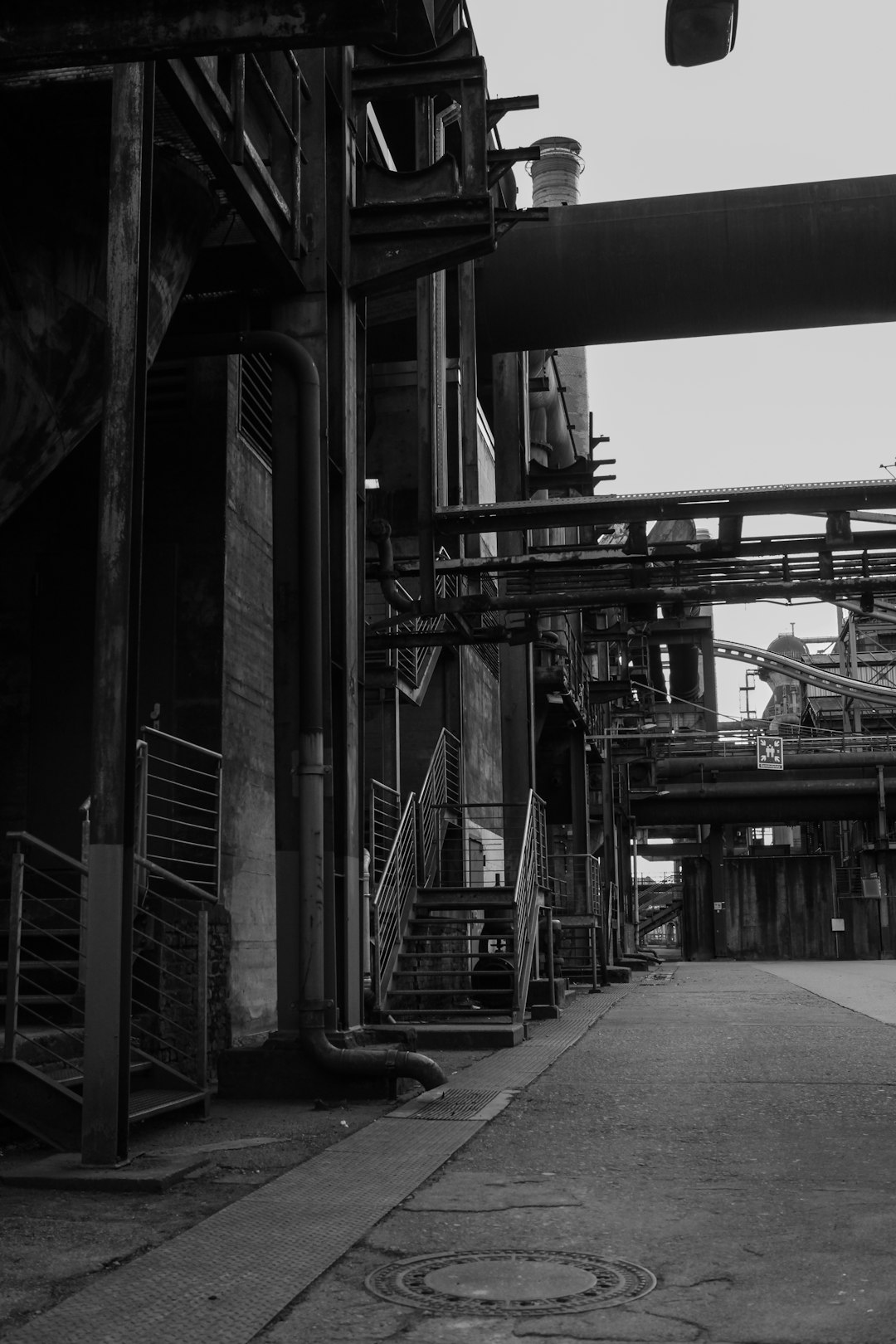 The industrial area of the city, black and white photography, wide angle shot, steel mill buildings, metal stairs leading to the factory entrance doors, industrial architecture, concrete walkways with pipes along side walls, distant machinery. –ar 85:128