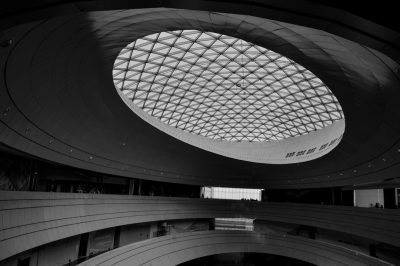 A black and white photo of the oval shaped glass roof inside an art museum in China, architecture photography, high angle shot, the circular skylight is covered in a geometric grid pattern, the building has three floors with large open space on the first floor for people to walk around, there are some crowds walking through it, the building walls have big digital screens displaying artwork, the overall style of composition resembles the style of [Kengo Kuma](https://goo.gl/search?artist%20Kengo%20Kuma). --ar 128:85