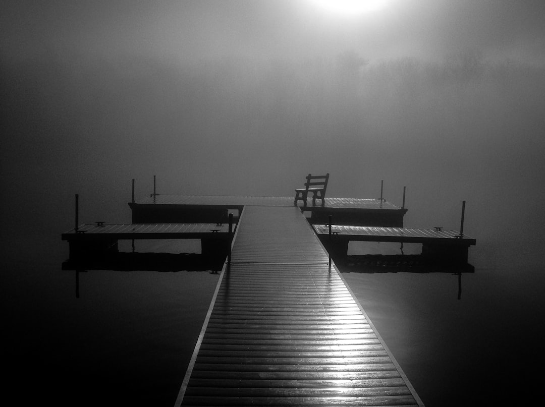 A black and white photo of a dock with a chair at the end, on a foggy night, beside a lake. The photo has the style of an artist known for their moody nighttime landscapes. –ar 128:95