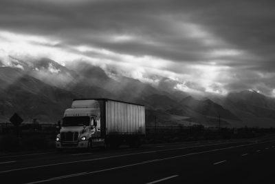 A black and white photo of a semi truck with a cargo trailer driving on a highway in front of mountains, with dark clouds above the mountains and light coming from behind. --ar 128:85