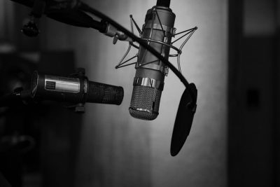 Black and white photo of an audio studio with microphones, focusing on the closeup view of one microphone suspended from above in the style of its stand. The background is a dark grey, creating contrast between light and shadow. The scene conveys the professional atmosphere of podcasting or recording music in a small space. --ar 128:85