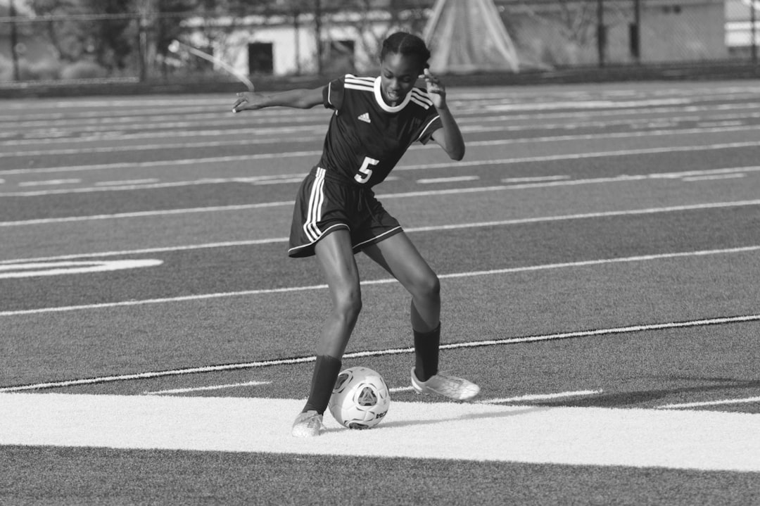 black and white photo of an African American high school soccer player, wearing black uniform shorts with the number “5” on them, playing in a game at her home field stadium, kicking a ball to start play or celebrating a goal, taken in the style of a Sony Alpha A7 III camera for a sports magazine cover. The photograph should capture sharp details and vibrant colors, highlighting the action as she skillfully handles the ball. The scene is set against a background of team banners and fans cheering from their seats in the bleachers, with clear skies above. –ar 128:85