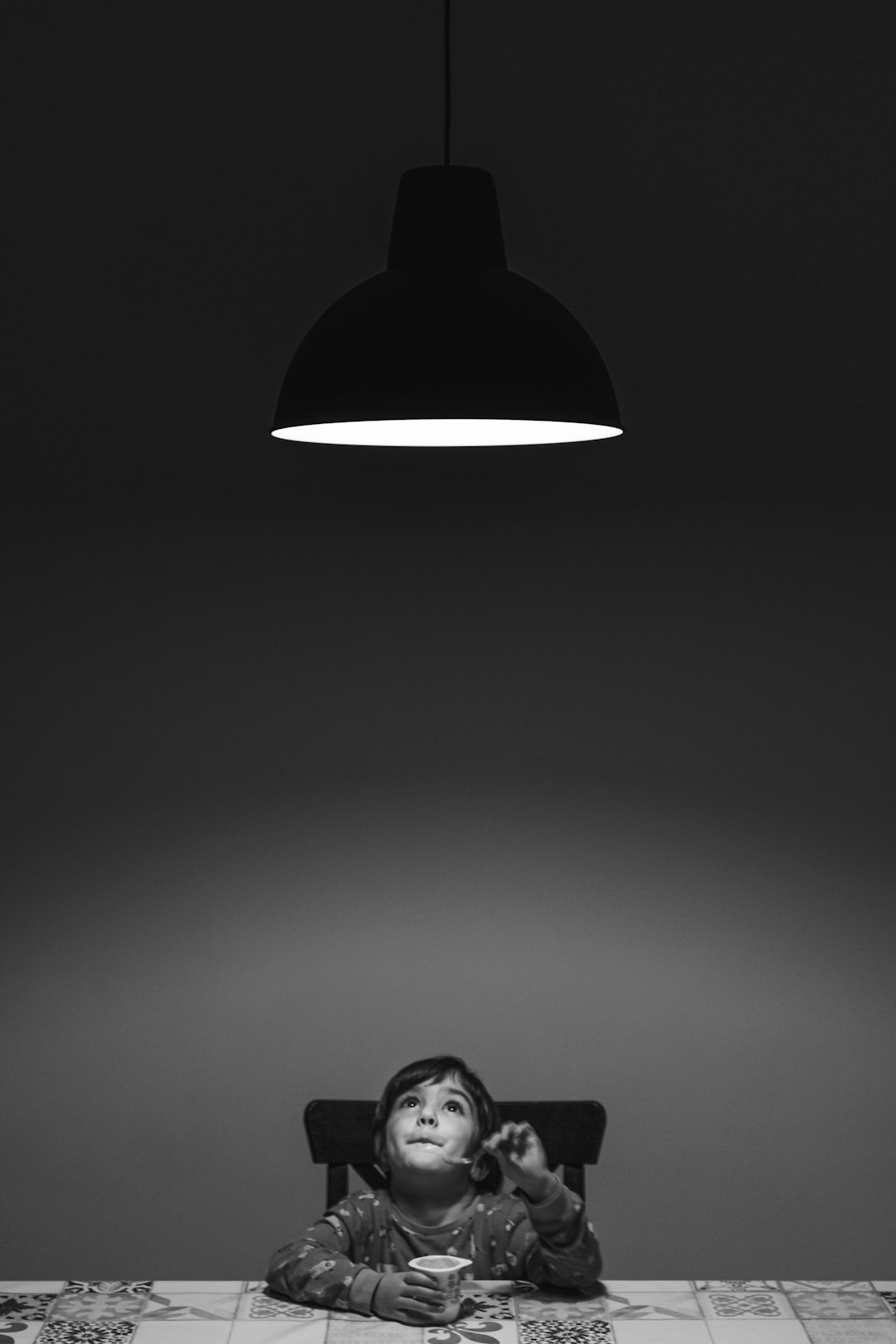 A child sitting at the dining table, looking up to see an overhead light hanging from above their head. The room is dark and the only source of illumination comes from that ceiling lamp. black white, high contrast, portrait photography, canon eos r5 –ar 85:128