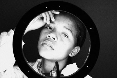 A black and white photo of an African American boy, looking at himself in the mirror with his hand on top of head. He has shaved hair. The camera is focused on him from above, capturing only half her face visible through circular frame. In front there's makeup, powder bl Extracted text, high contrast. Shot by arriflex 35 BL Camera Canon K35 Prime Lenses, medium shot, Kodak Gold 200 film. --ar 128:85