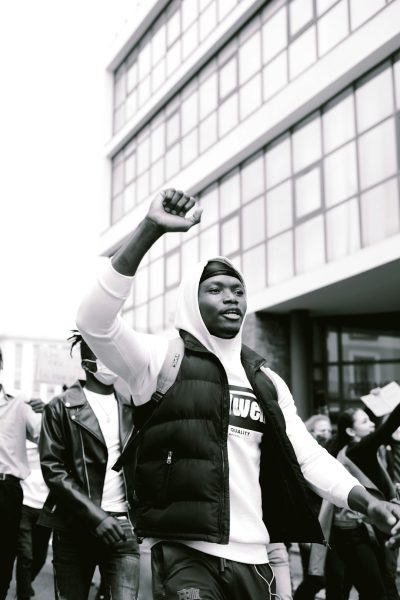 Black and white street photography of a black man in a puffer vest, holding up his fist while marching with a crowd outside an office building during a protest against police shootings of Black people, in the style of Arri Alexa 65mm camera, cinematic, low angle, symmetrical composition, color grade. --ar 85:128