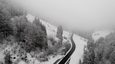 aerial view of black and white road in the mountains with snow, surrounded by forest trees, foggy weather, shot on Sony Alpha A7 III --ar 16:9