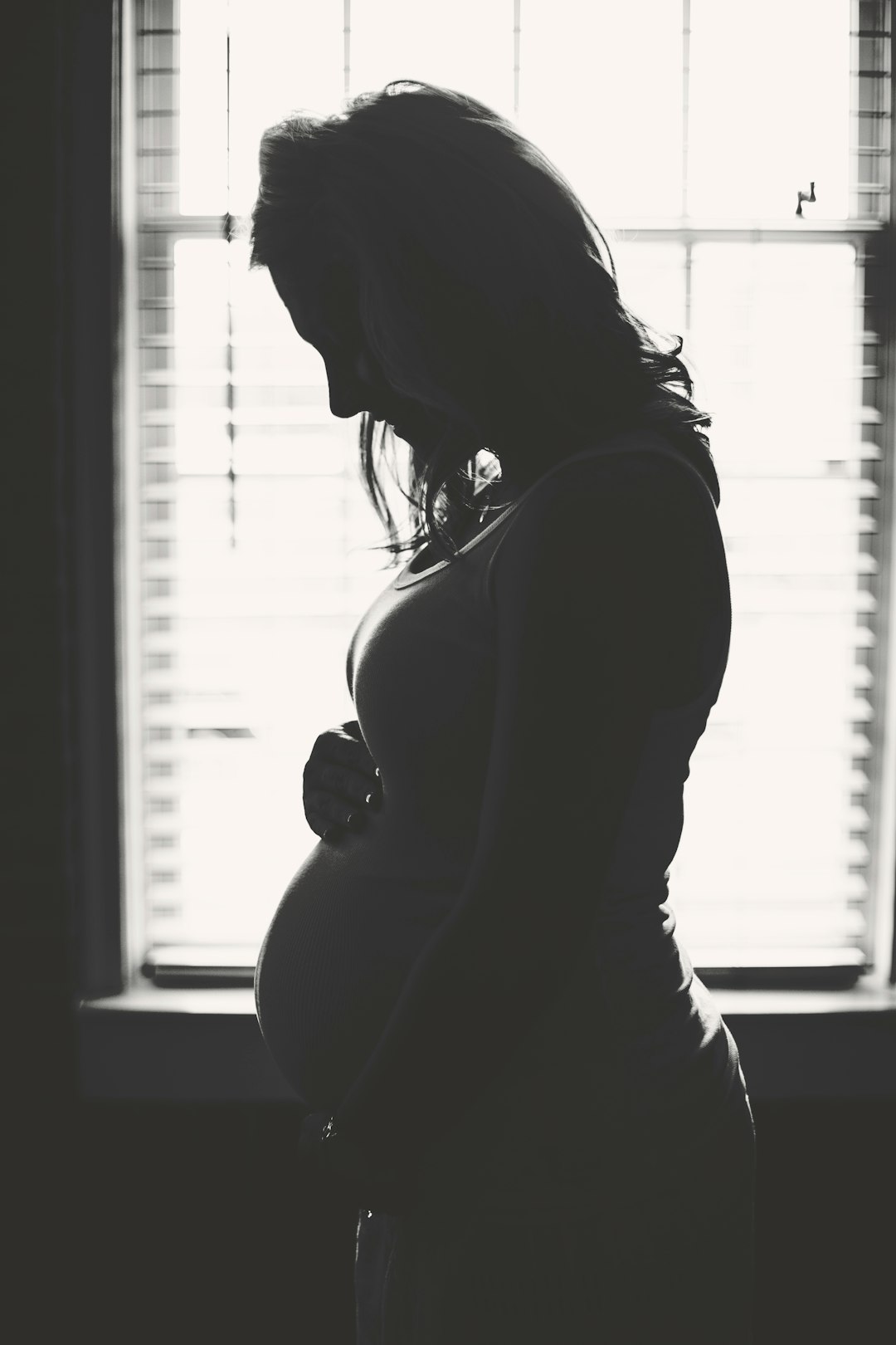 A pregnant woman’s silhouette against the soft light of an open window, her belly gracefully round and visible in black and white photography, creating a timeless and serene atmosphere for potential parents to gaze upon their future child. The image is in the style of soft light and graceful silhouettes. –ar 85:128