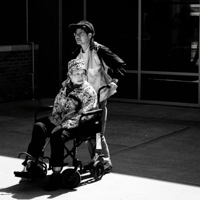 A young man in his thirties is pushing an elderly woman sitting in her wheelchair outside of a hospital. The scene is captured from behind with the sun shining down and casting shadows. The atmosphere looks somber, and the mood conveys serenity but melancholy. The composition has strong contrast between black and white tones. The photo was taken in the style of a Leica M6 camera using Fujifilm film.