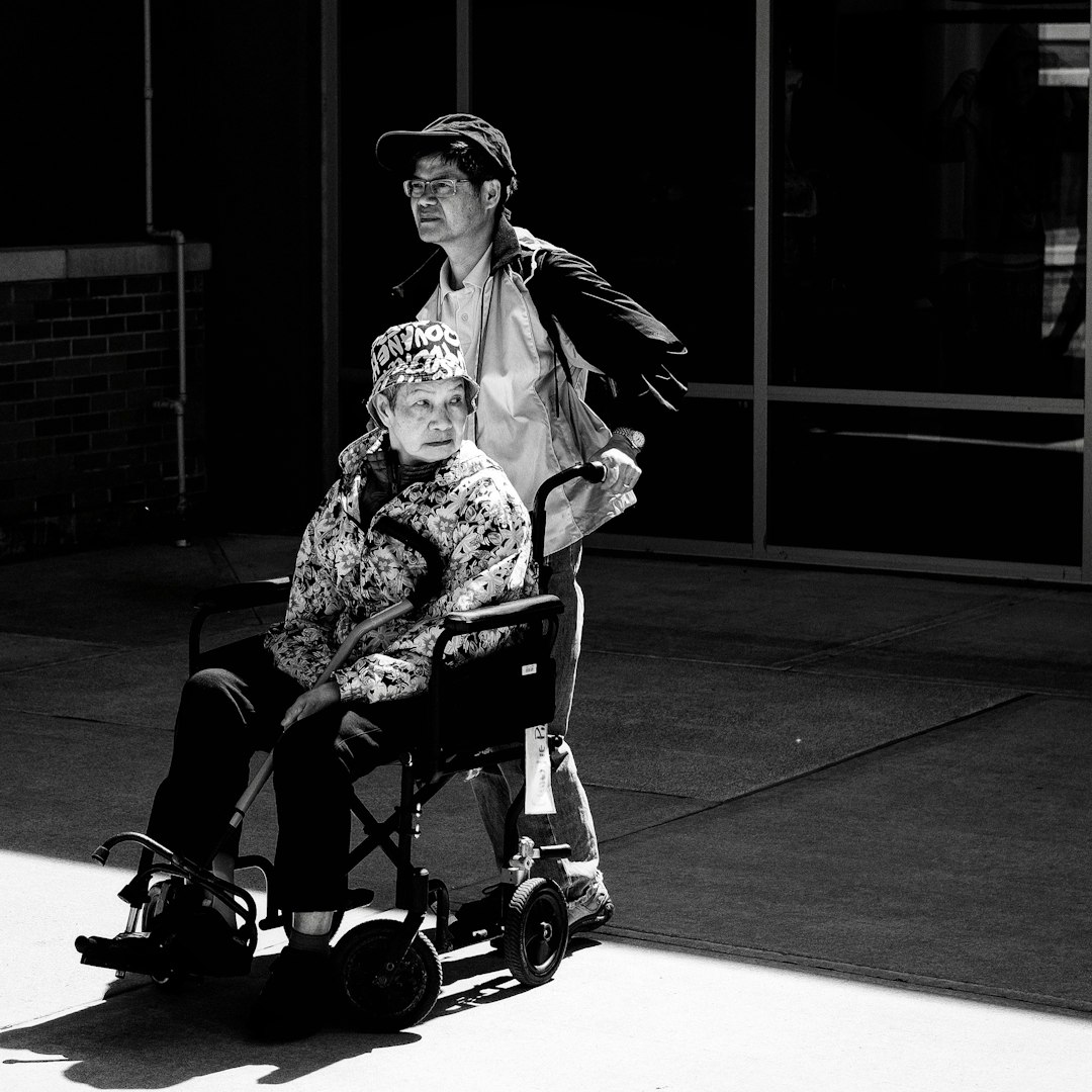 A young man in his thirties is pushing an elderly woman sitting in her wheelchair outside of a hospital. The scene is captured from behind with the sun shining down and casting shadows. The atmosphere looks somber, and the mood conveys serenity but melancholy. The composition has strong contrast between black and white tones. The photo was taken in the style of a Leica M6 camera using Fujifilm film.