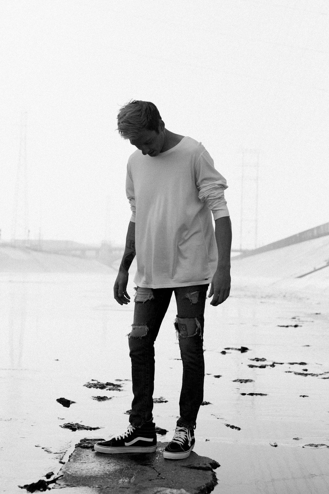 high contrast black and white photo of justin bickle in an oversized long sleeve, ripped jeans, standing on the beach with his feet in the water, wearing converse sneakers, holding a skateboard under one arm. Shot in the style of arriflex 35 BL Camera, Canon K35 Prime Lenses, 70mm. –ar 85:128