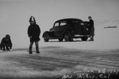 Old black and white photo of Inuit people on the tundra. One person is standing in front holding onto an old car that has washed up from the sea to land on snow with "Fuppy for Bishon" written on it. The other two people stand next to them looking at the camera. One man wears traditional  while another woman stands wearing a long coat and hat. It's winter and there are no trees or buildings around just flat ground covered in snow. --ar 128:85