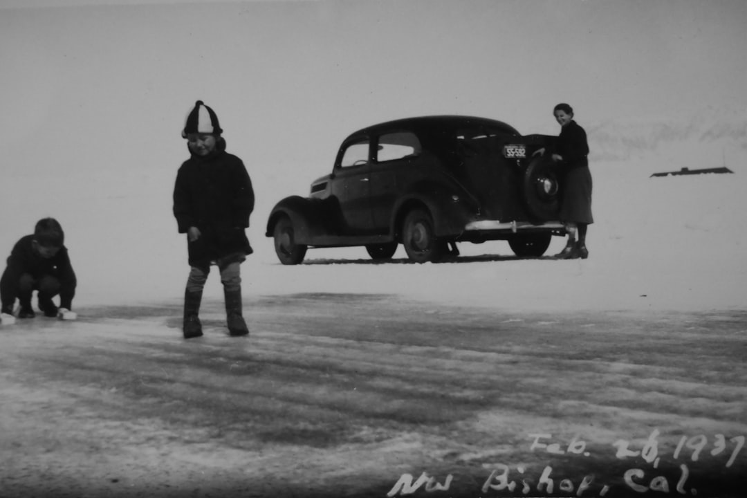 Old black and white photo of Inuit people on the tundra. One person is standing in front holding onto an old car that has washed up from the sea to land on snow with “Fuppy for Bishon” written on it. The other two people stand next to them looking at the camera. One man wears traditional  while another woman stands wearing a long coat and hat. It’s winter and there are no trees or buildings around just flat ground covered in snow. –ar 128:85