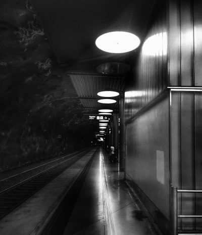 A black and white photo of the underground station in Stockholm, Sweden. The picture was taken with an iPhone camera. It's night time. There is only one light on at ground level, and it illuminates everything around the train tracks. A few people can be seen walking. The wall to your right has large metal panels. In the background there's a dark tunnel that leads under water. On top of some walls there are flat led lights, but they are not lit up yet. --ar 55:64