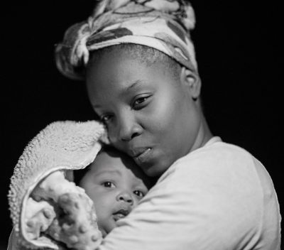 A black and white portrait of an African American mother holding her newborn baby, illuminated by soft light against the dark background. The woman has short hair with blonde highlights, wearing a head bandana. She is looking down at the camera with affectionate eyes that reflect love and joy as she cradles the child close to their chest. This photo was taken using a Fujifilm XT4 camera with an aperture f/2 lens set on a focal length macro, in the style of the photographer. --ar 8:7