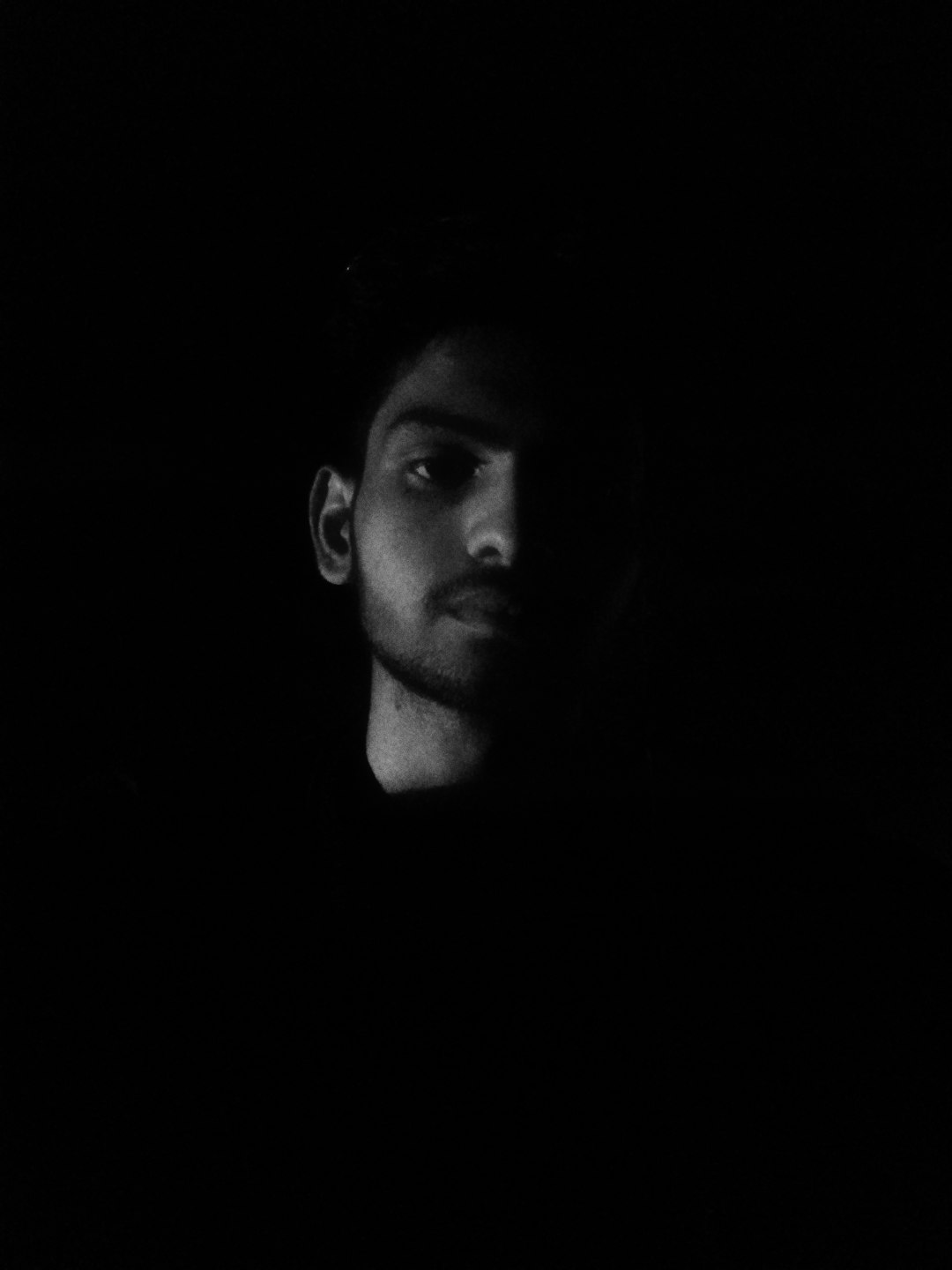 a black and white portrait of an indian man in the dark, he is just slightly visible due to lighting, his face has some shadow on it, he looks confident but serious, he’s wearing tshirt, shot with canon eos r5 –ar 3:4