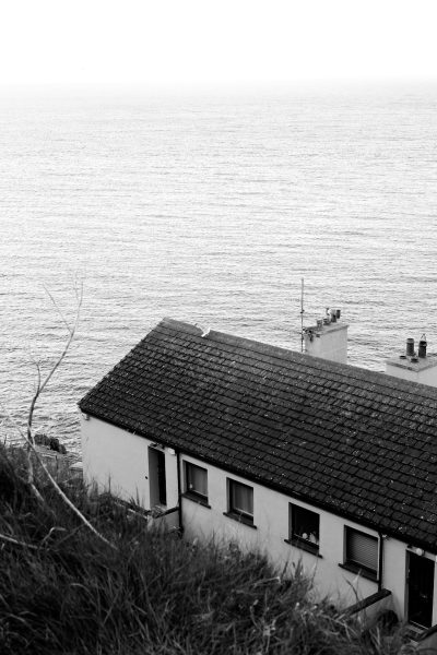 A black and white photograph of an Irish cottage with the sea in the background, taken from above in the grainy film noir style with high contrast and overcast lighting. The photograph was shot on a Hasselblad medium format camera using Kodak Portra 400T film. --ar 85:128