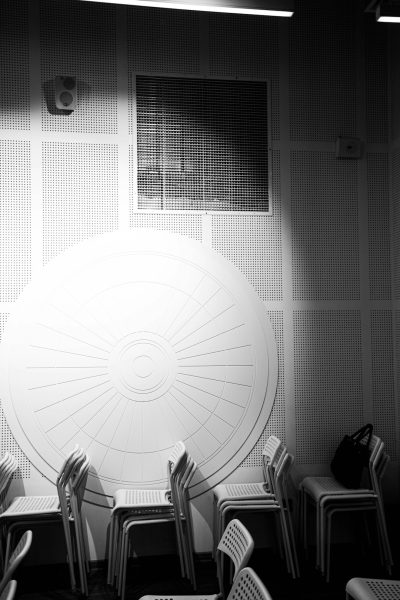 A black and white photograph shows the interior wall of a room with chairs. A large round graphic depicting a darts board is on the wall. The floor is covered with stackable plastic chairs. The room has a window at one end. An industrial light hanging from the ceiling illuminates the scene. The walls have small holes covering parts or all of them with white dots. --ar 85:128